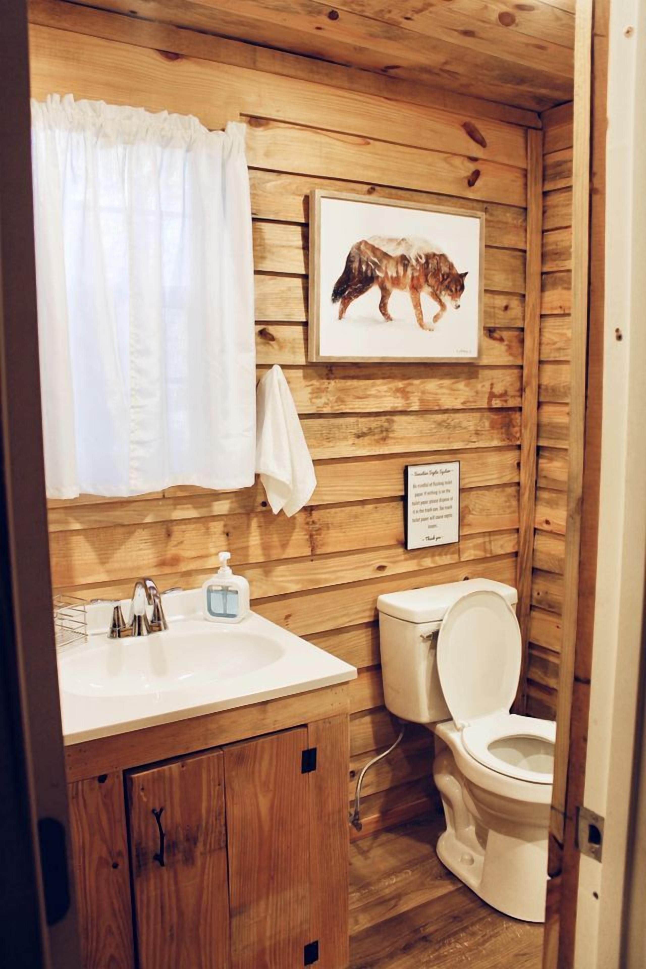 Une salle de bain rustique, aménagée dans une cabane en bois, avec un lavabo, des toilettes et un tableau représentant un loup accroché au mur.