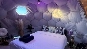 A hexagonal tent-style room with a bed, pillows, a small table, and a hanging light fixture.