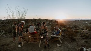 Un gruppo di persone all'aperto, alcune sedute su panchine, altre in piedi, in un ambiente desertico con scarsa vegetazione e montagne in lontananza.