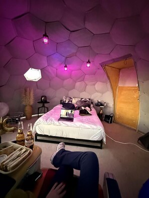 A room with a hexagonal ceiling, a bed with a laptop, a chair, and a table with bottles. - Unique Dome with Great Desert Views in Terlingua, Texas (Terlingua)