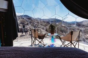 Una habitación con un separador de vidrio hexagonal, dos sillas, una mesita y una planta en maceta.