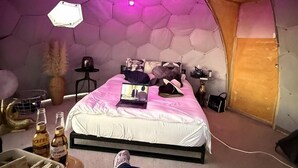 A room with a hexagonal ceiling, a bed with a laptop, a chair, and a table with bottles.