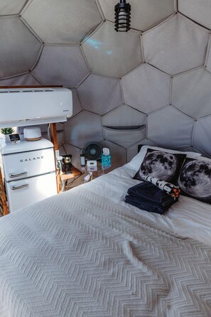 A bedroom with a hexagonal headboard, a bed with a white herringbone-patterned bedspread, a bedside table with a fan, and a small cabinet labeled 'GALANZ'.