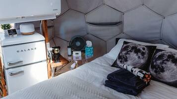 A bedroom with a hexagonal headboard, a bed with a white herringbone-patterned bedspread, a bedside table with a fan, and a small cabinet labeled 'GALANZ'.