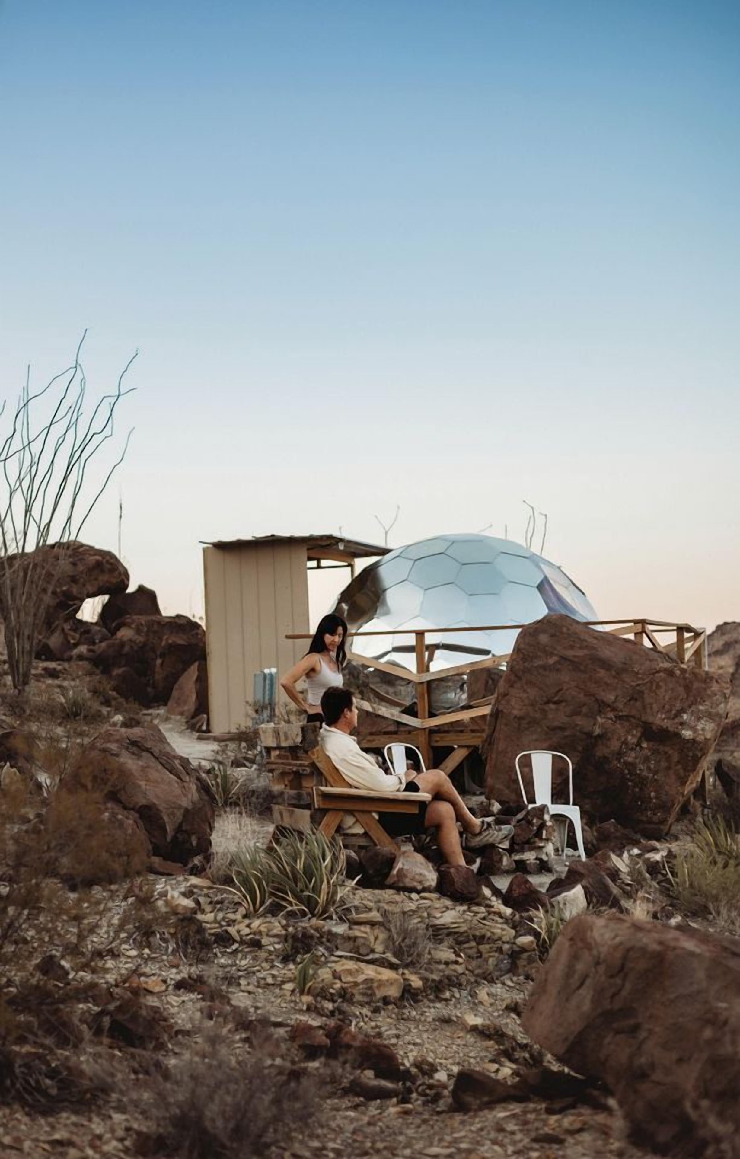A rustic outdoor setting with a large transparent dome structure, wooden seating, and a small wooden shelter.