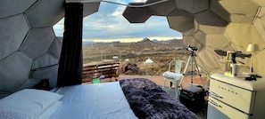 A bedroom with a bed, a dresser, a tripod, and a view of a desert landscape.
