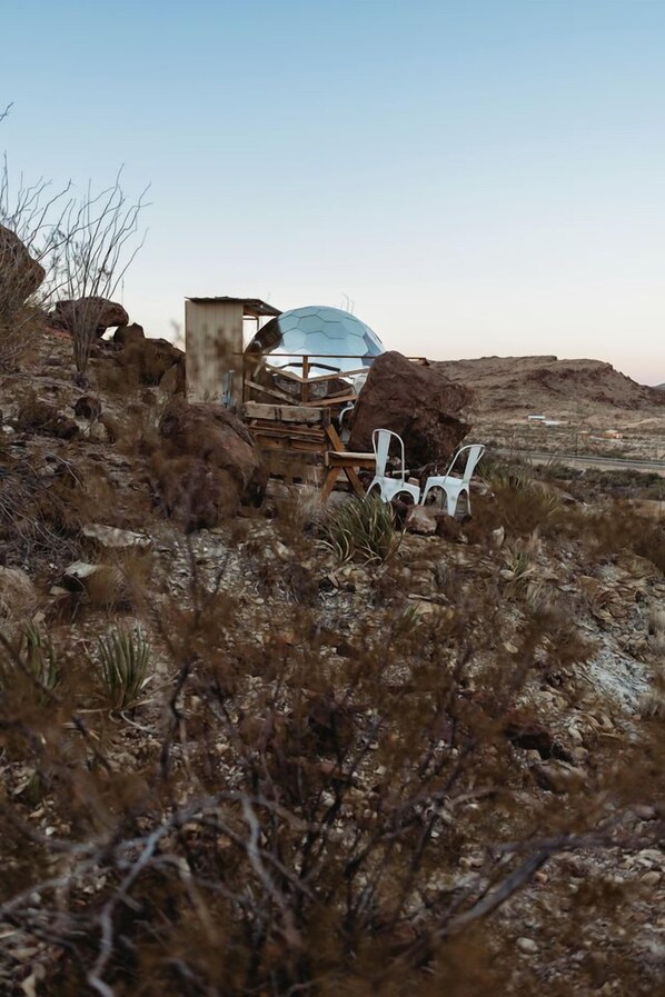 Un paysage désertique avec une grande structure en forme de dôme transparent, des chaises en plastique blanc et un petit abri en bois.