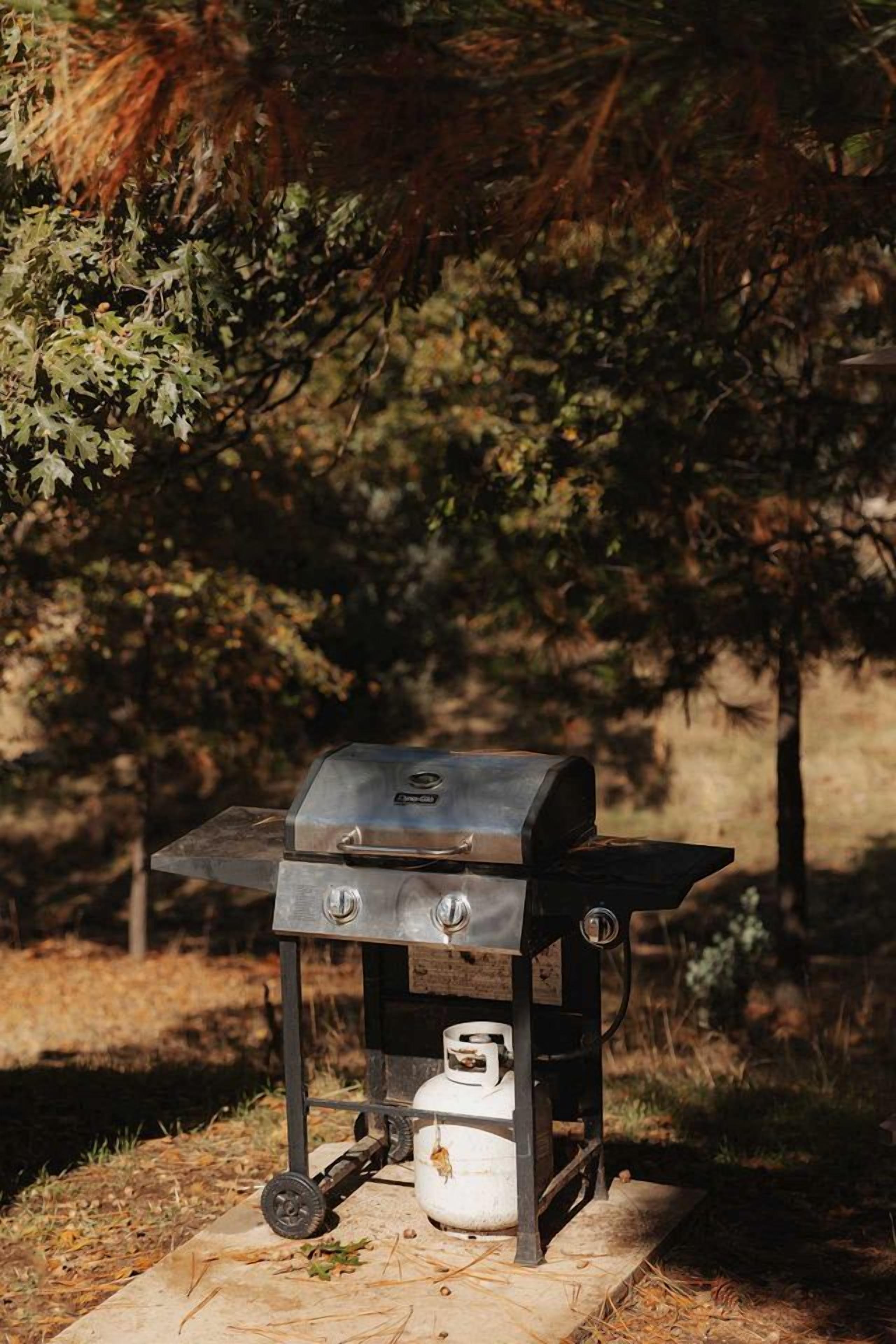 Een draagbare barbecue met gasfles en wielen, geplaatst op een betonnen ondergrond in een bebost gebied.