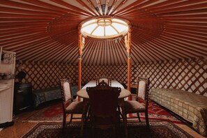 A traditional yurt interior with a central round table, four chairs, a bed, and a patterned rug.