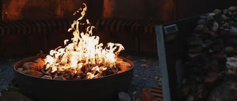 A fire pit with burning logs, surrounded by rocks and gravel, with a building in the background.