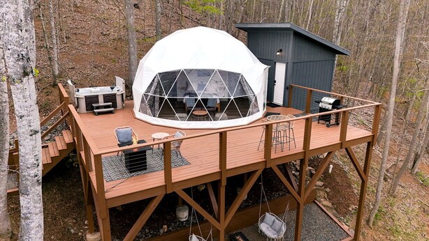 Une structure en forme de dôme géodésique, dotée d’une terrasse en bois, d’un espace salon extérieur et d’un petit bâtiment.
