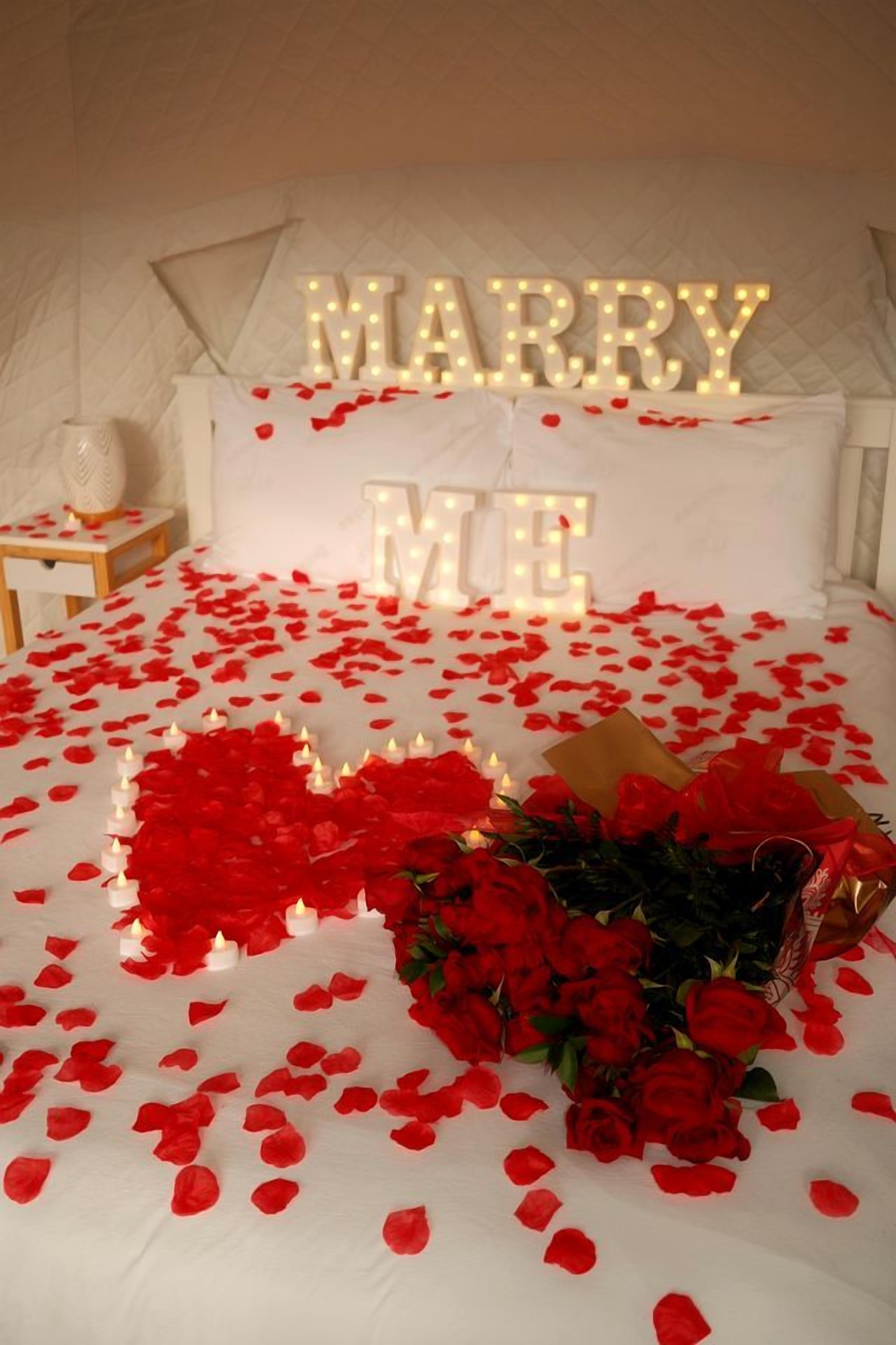 A bed with 'MARRY ME' illuminated letters, red rose petals, and a bouquet of roses.