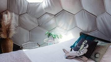 A room with a hexagonal patterned headboard, a bed with white bedding, a hanging lamp, and a small plant.