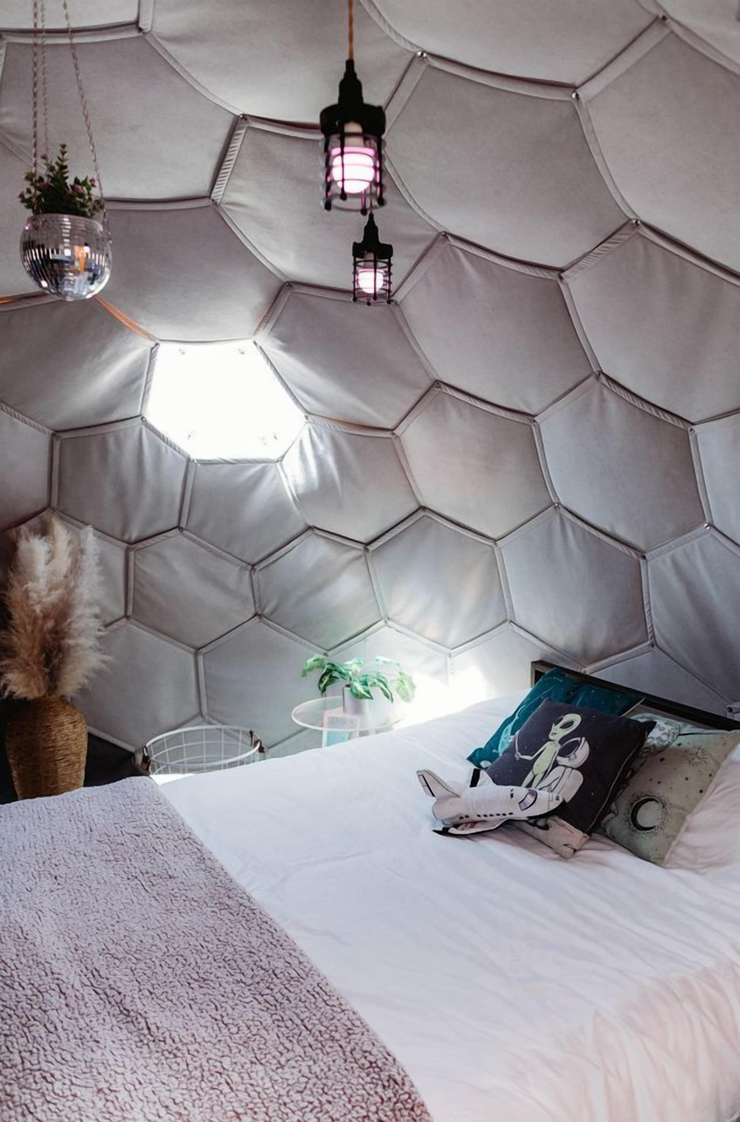 Una habitación con tablero de cama de diseño hexagonal, ropa de cama blanca, una lámpara colgante y una planta pequeña.