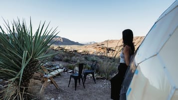 A desert landscape with a person standing near a tent and chairs, with mountains in the background.