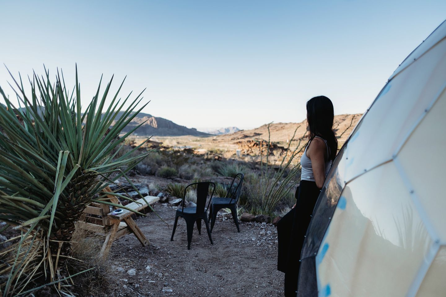 Un paysage désertique avec une personne debout près d’une tente et de quelques chaises, des montagnes se dressant à l’arrière-plan.