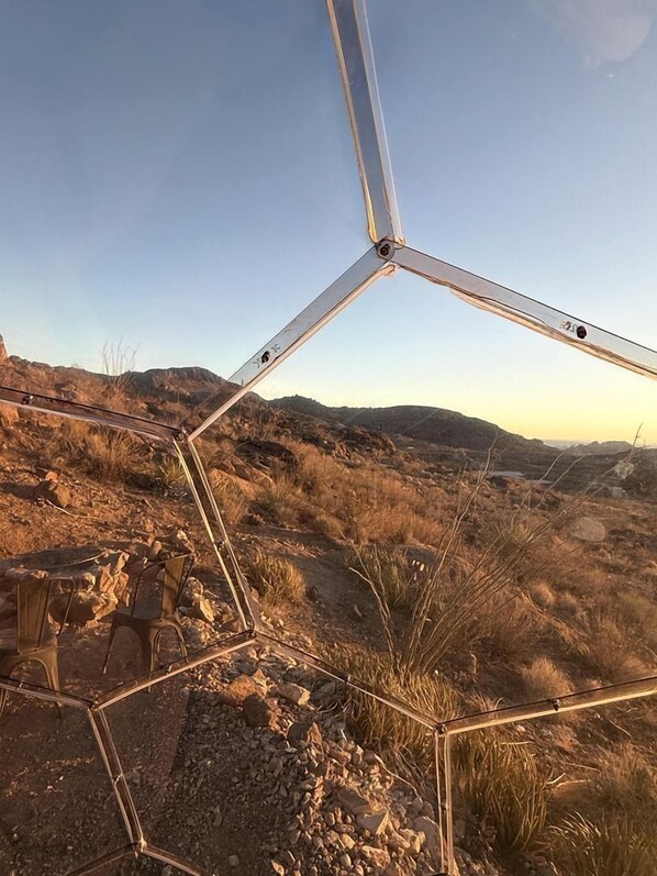 A geometric metal structure in a dry, rocky landscape with distant hills.