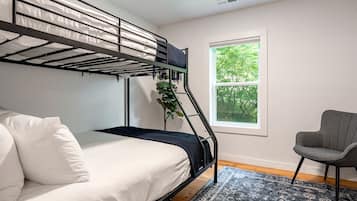 A bunk bed with a mattress and pillows, a chair, a window with a view of trees, and a patterned rug.