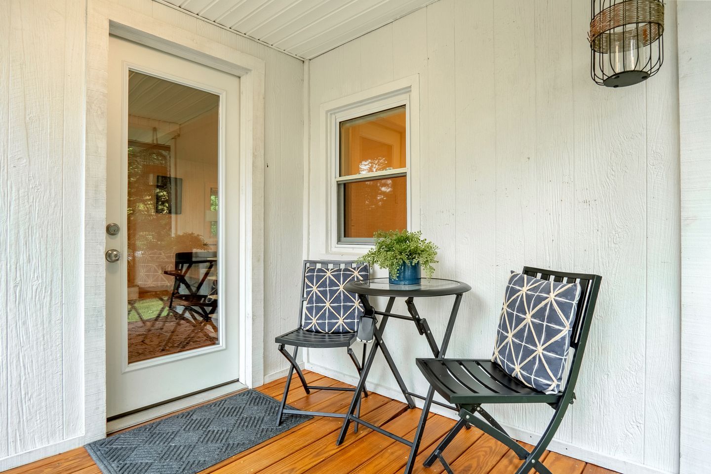 A porch with a door, two chairs, a small table, a potted plant, and a hanging light fixture.