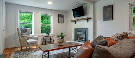 A living room with a fireplace, a television mounted on the wall, and a large window with a view of greenery.