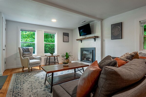 A living room with a fireplace, a television mounted on the wall, and a large window with a view of greenery.