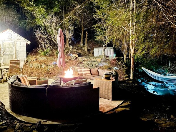 A stone patio with outdoor furniture, a fire pit, and a canoe by a stream.