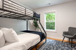 A bunk bed with a mattress and pillows, a chair, a window with a view of trees, and a patterned rug.