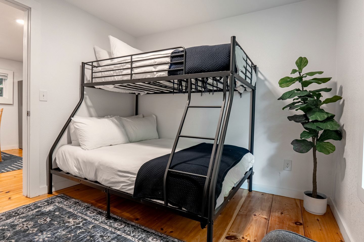 Un lit superposé avec une échelle, du linge de lit blanc, une couverture foncée, un tapis à motifs et une plante en pot.
