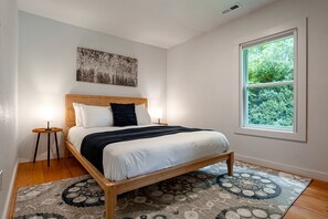 Un dormitorio con cama de madera, ropa de cama blanca, un plaid oscuro, una mesita de noche con lámpara, un ventanal con vistas a zonas verdes y una obra de arte colgada en la pared.