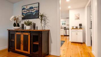 A hallway with a wooden floor, a dark wooden cabinet with glass doors, a painting on the wall, and a kitchen area in the background.