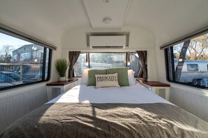 Ein Schlafzimmer mit Bett, Kissen, einem Fenster, Vorhängen, einer Pflanze und Blick ins Freie.