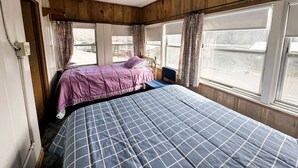 Una habitación con dos camas, paredes de madera, ventanas amplias con cortinas y una puerta visible.