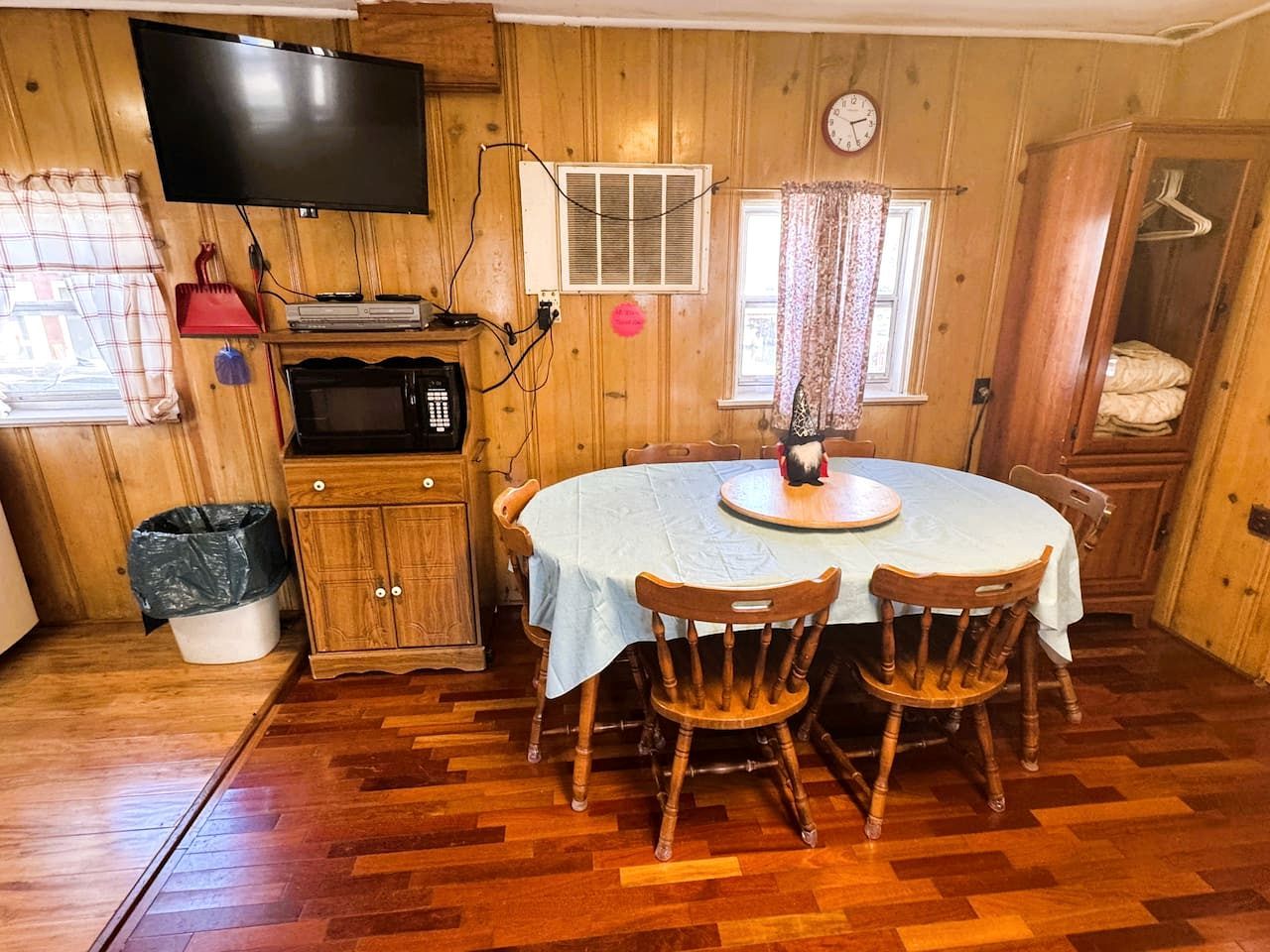 Een kamer met houten lambrisering, een eettafel, stoelen, een televisie, een magnetron en een klok aan de muur.