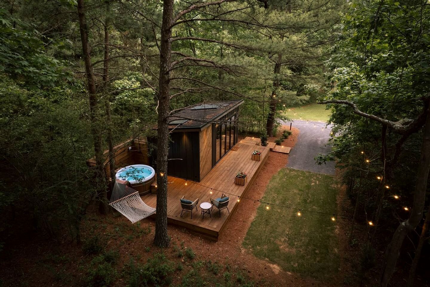 A wooden deck with outdoor seating, a hot tub, and a small building, surrounded by dense forest.