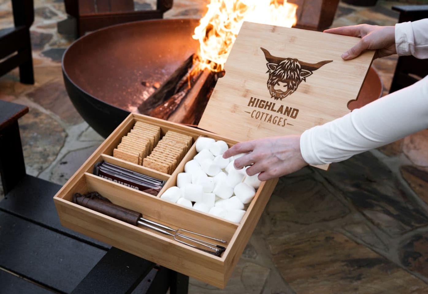 A wooden box with a logo and text, held by a person, next to a fire pit with logs.