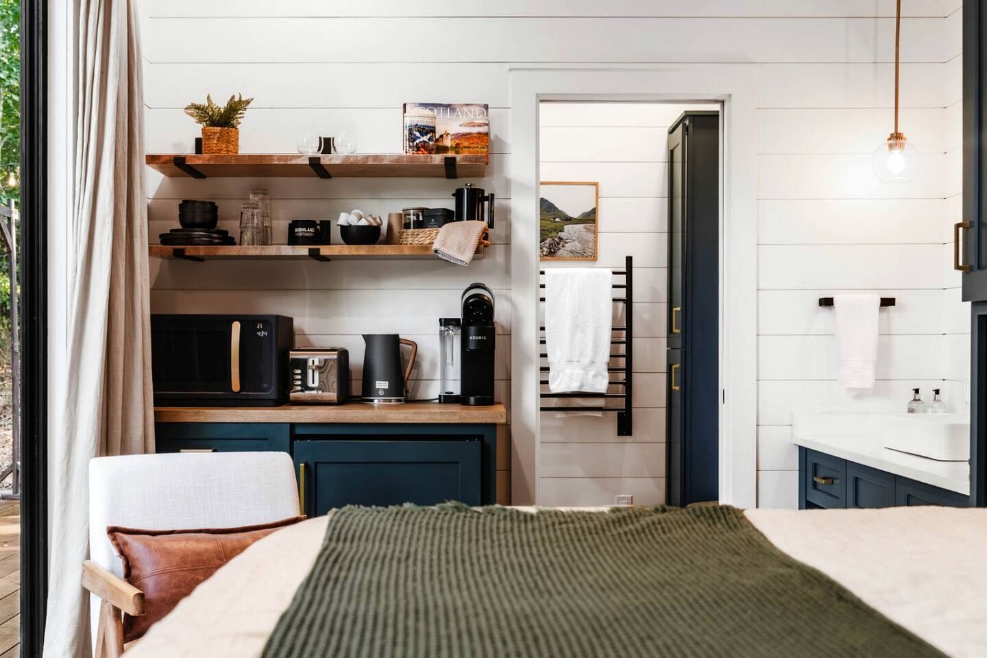 A compact room with a bed, a desk, a coffee machine, a kettle, and a shelf with books and decorative items.