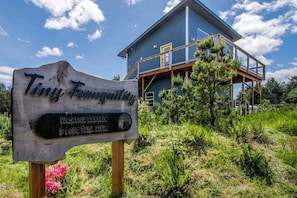 A wooden sign with 'Tiny Tranquility' and 'VICATION RENTALS & LONG TERM SITES' text, a blue building with a balcony, and surrounding greenery.
