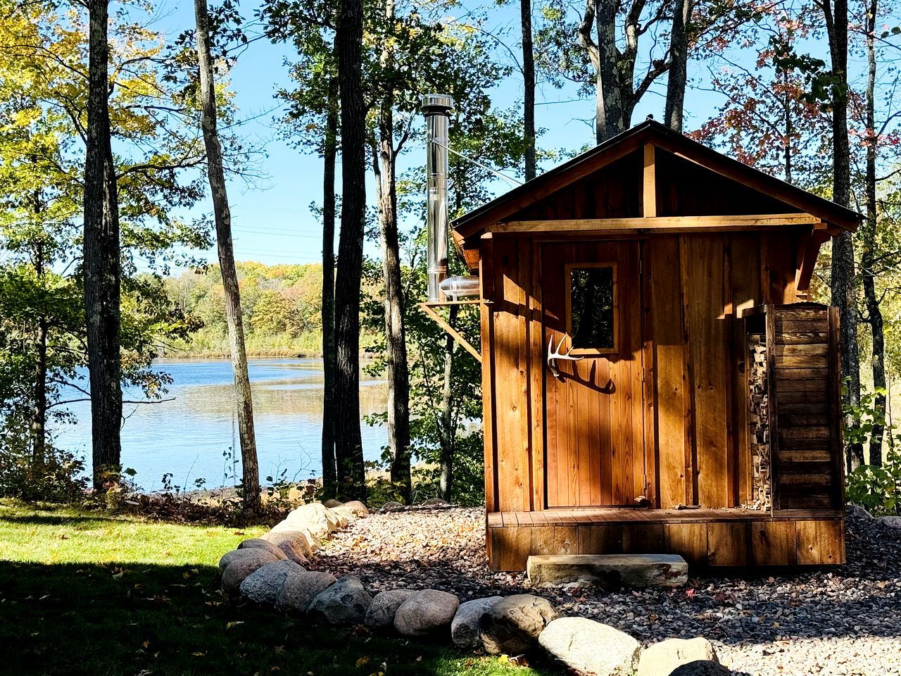 A wooden cabin with a chimney, situated by a lake surrounded by trees.