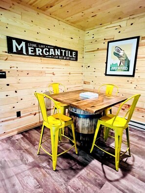 Un intérieur en bois avec une table et quatre chaises jaunes, une enseigne indiquant « STONE LAKE MERGANTILE, appâts vivants et produits secs », et un tableau encadré représentant une baleine.