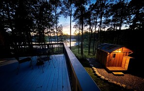 A wooden deck with a table and chairs, overlooking a forest and a body of water at sunset. - Pet-Friendly Lakeside Cabin Ideal for a Family Getaway in Wisconsin (Stone Lake)