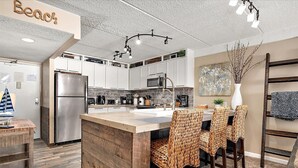 A modern kitchen with white cabinets, a stainless steel refrigerator, and a central island with woven chairs. - Direct Oceanfront Two Bedroom Condo with Pool (Ocean City)