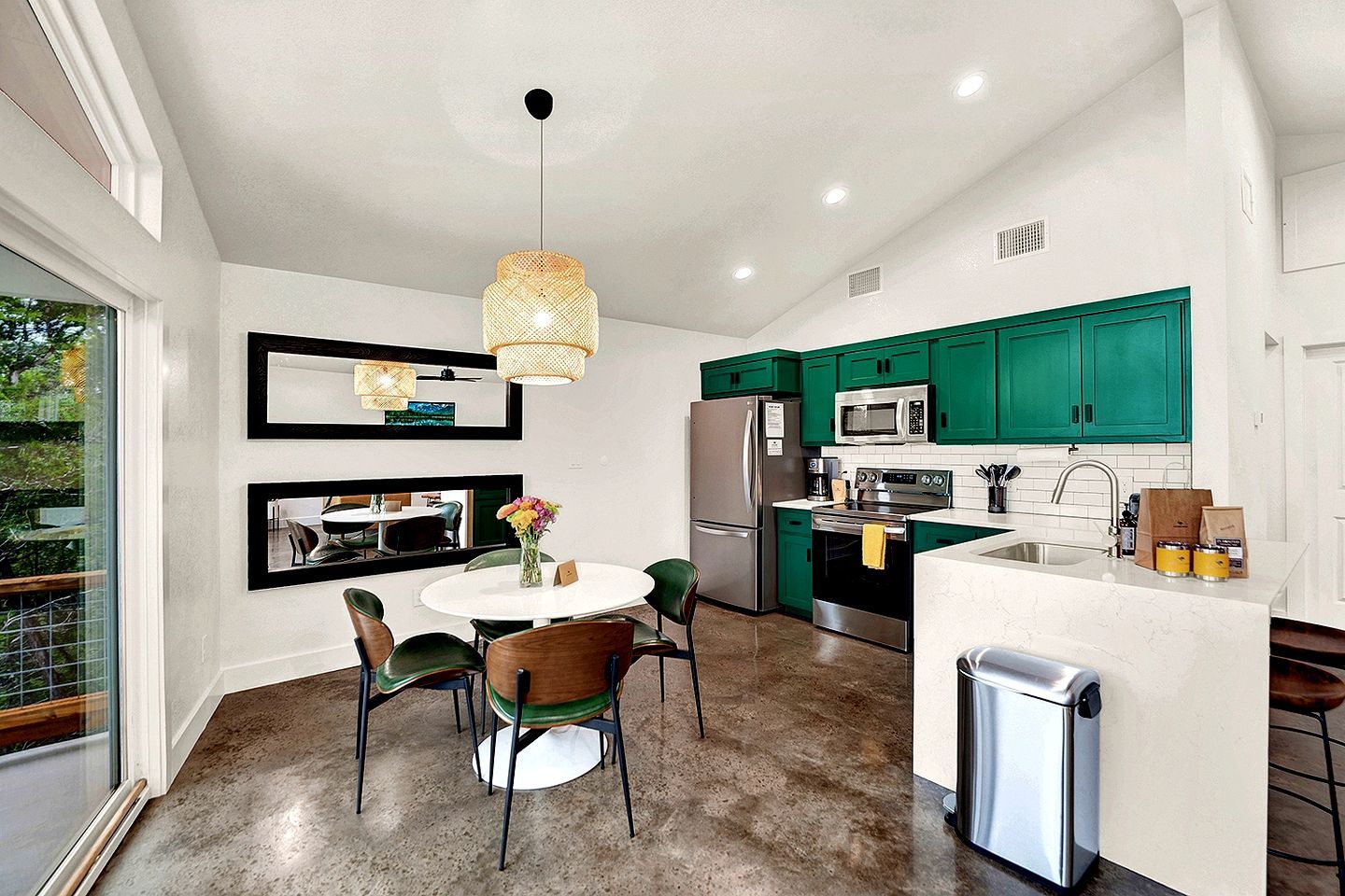A modern kitchen with green cabinets, stainless steel appliances, a round dining table with chairs, and a hanging light fixture.