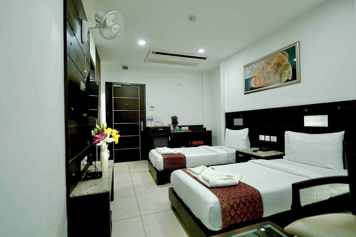 Habitación de hotel con dos camas, un escritorio, una silla, un televisor, un ventilador y un cuadro en la pared.