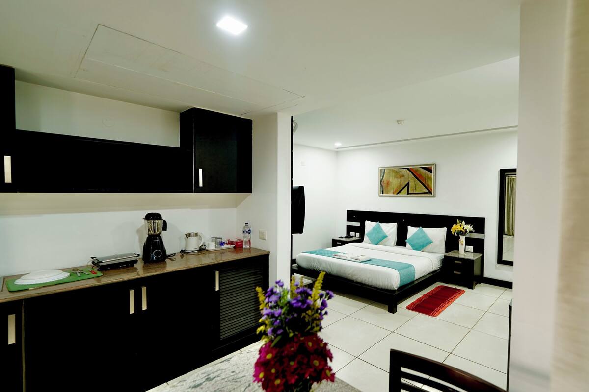 Habitación de hotel con kitchenette, cama con cubrecama rojo y blanco, un cuadro en la pared y un florero con flores en la mesita de noche.
