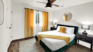 Habitación de hotel con cama, ventilador de techo, ventana con cortinas, mesita de noche con lámpara y espejo en la pared.