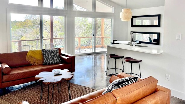 Amplia sala de estar con un gran ventanal, ventilador de techo, sofá, mesa de centro y área de cocina con taburete de bar.