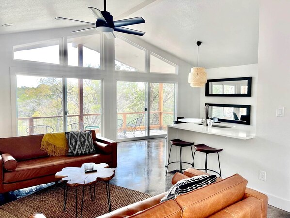 A spacious living area with a large window, a ceiling fan, a sofa, a coffee table, and a kitchen area with a bar stool. - Splendid Chic Cabin with Modern Interior and Pool in Wimberley, Texas (Wimberley)