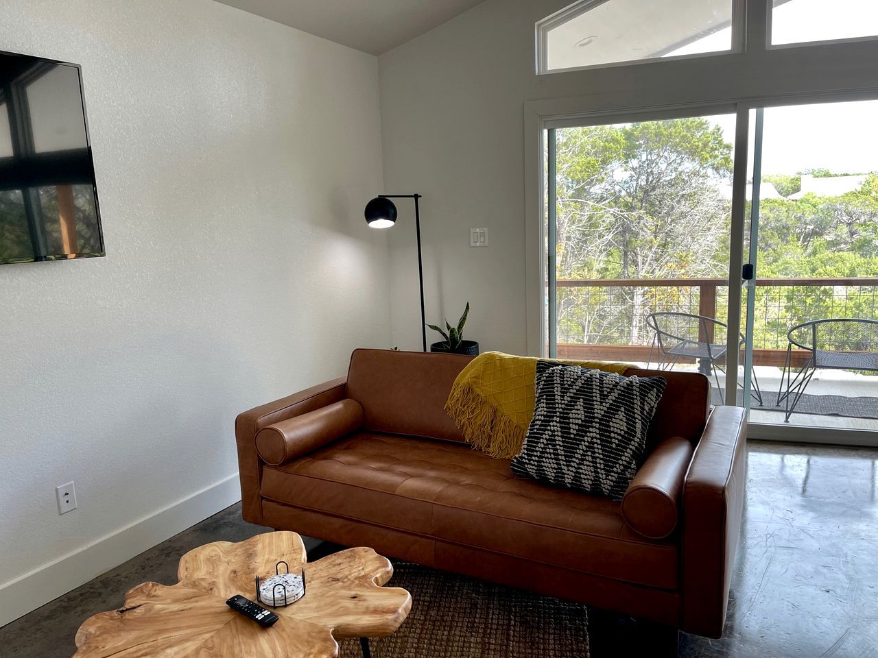 Un moderno salón con un sofá de cuero marrón, una mesa de centro de madera, una lámpara de pie y vistas al exterior a través de un ventanal.