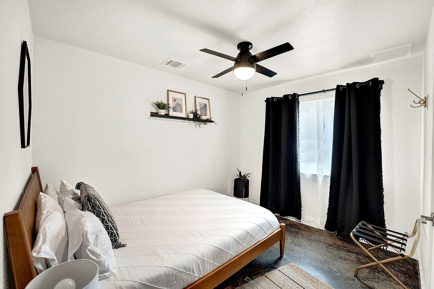A bedroom with a bed, a ceiling fan, a window with curtains, a small plant, and a wall-mounted shelf.