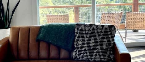 Um sofá de couro marrom com uma manta verde e uma almofada estampada, uma caneca branca sobre um apoio de madeira, e uma varanda com cadeiras de vime com vista para uma paisagem arborizada.